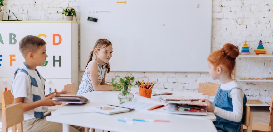 Three primary school children sit at a table in front of a whiteboard in a classroom.